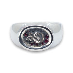 May include: A silver ring with an oval-shaped design featuring a stylised rat with red gemstones around the edge.