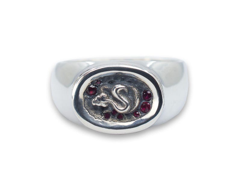 May include: A silver ring with an oval-shaped design featuring a stylised rat with red gemstones around the edge.