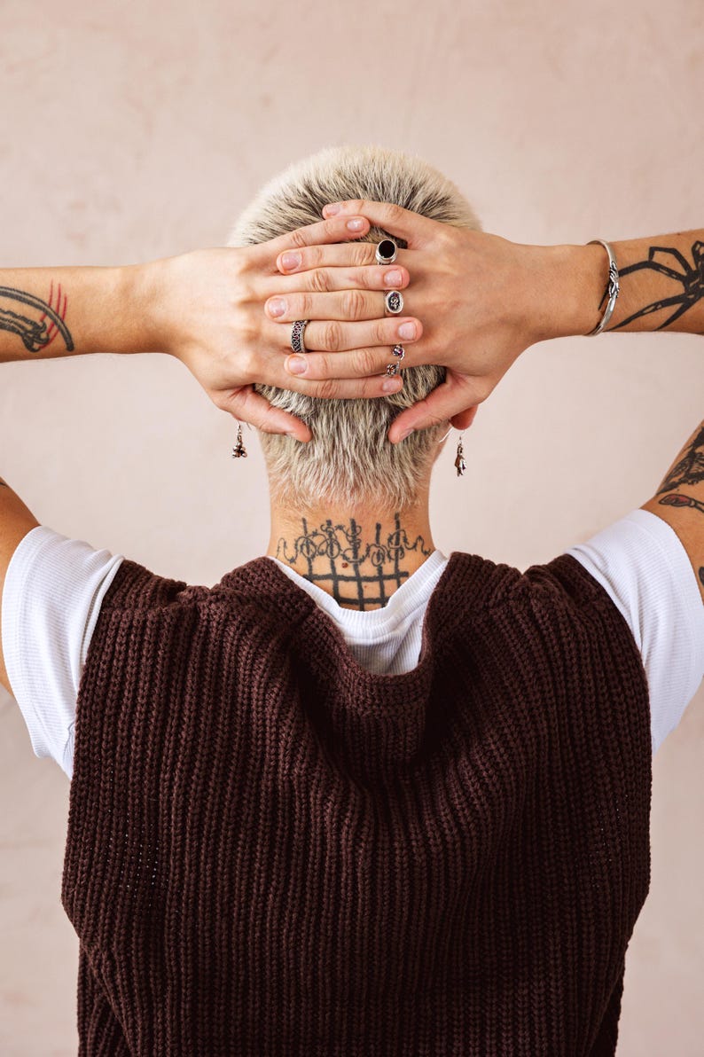 May include: A person with short, blonde hair wearing a brown knitted waistcoat over a white t-shirt. They have multiple silver rings on their fingers and a tattoo on their neck.