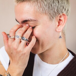 May include: A person wearing a white t-shirt and a brown cardigan with a silver necklace and a silver bracelet. They are wearing several silver rings on their fingers and a pair of silver earrings with purple stones.