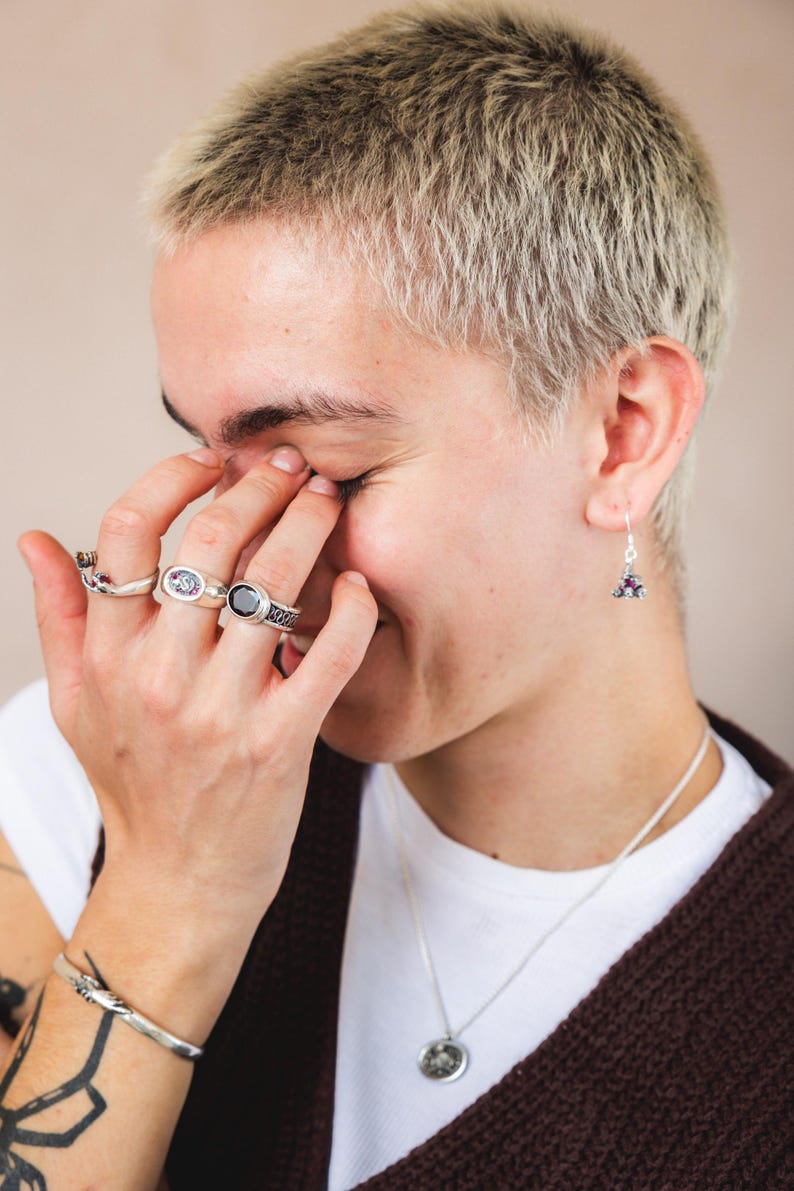 May include: A person wearing a white t-shirt and a brown cardigan with a silver necklace and a silver bracelet. They are wearing several silver rings on their fingers and a pair of silver earrings with purple stones.