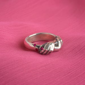 May include: A silver ring with two hands clasped together, adorned with small pink gemstones.