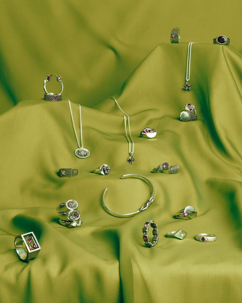 May include: A collection of silver rings and necklaces with red gemstones displayed on a green fabric background. The jewellery features a variety of designs, including a ring with a small crown, a ring with a cross, and a ring with a heart.