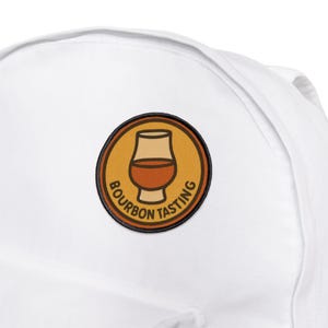 May include: White backpack with a circular patch featuring a bourbon glass graphic. The patch has a brown and tan color scheme with the words "Bourbon Tasting" in a circular design around the glass.