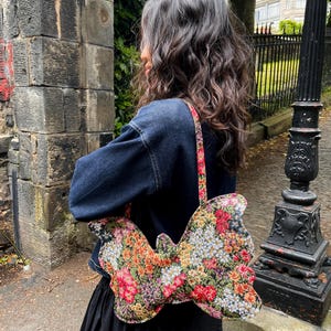 May include: A floral butterfly-shaped handbag with a colorful pattern of red, orange, yellow, and white flowers. The bag has a matching strap and is carried over the shoulder. The person is wearing a dark blue denim jacket and a black skirt.