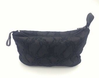 Op de afbeelding: Een zwart tasje met ritssluiting en een buitenlaag van delicate kanten stof met bloemmotief. Het accessoire is voorzien van een zwarte ritssluiting met aan beide uiteinden een klein treklipje voor het hoofdvak van de tas.