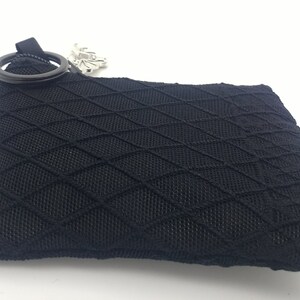 May include: A black rectangular pouch featuring a raised diamond-shaped grid pattern across its surface. The pouch includes a black circular ring attached to the top corner and a small silver-toned metallic charm dangling from the zipper pull.