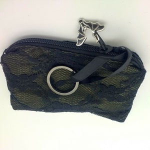 May include: A small black zippered pouch featuring an olive green fabric base overlaid with black floral lace. The zipper pull is a silver-toned metal bat charm, and a black fabric strap with a circular metal key ring is attached to the zipper.