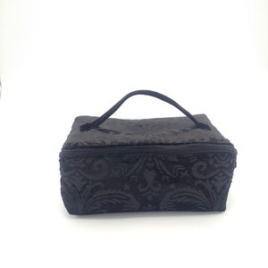 May include: A rectangular black cosmetic bag featuring a textured damask pattern across its surface. The bag includes a single black carrying handle attached to the top and a zippered closure that runs along the upper perimeter of the structure.