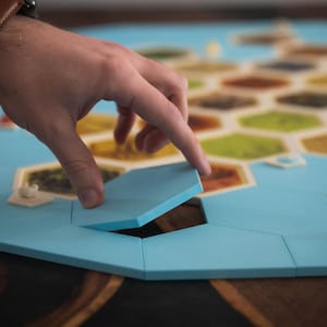 Könnte beinhalten: Ein Brettspiel mit einem hellblauen Spielbrett und bunten sechseckigen Kacheln. Eine Hand legt eine hellblaue Kachel in das Brett. Das Spielbrett hat kleine weiße und braune Spielsteine.