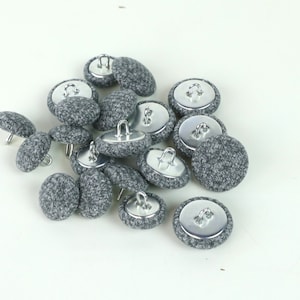 Puede incluir: Un montón de botones cubiertos de tela gris con respaldos y bucles de metal plateado. Los botones son todos del mismo tamaño y forma.