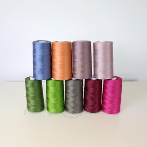 Op de afbeelding: Een verzameling van tien klossen garen in verschillende kleuren, waaronder blauw, oranje, grijs en roze. De klossen zijn in twee rijen gerangschikt, met de bovenste rij met vier klossen en de onderste rij met zes.