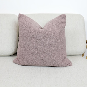 May include: A soft, plush cushion with a mauve colour and a fuzzy texture. The cushion is sitting on a light-coloured sofa.