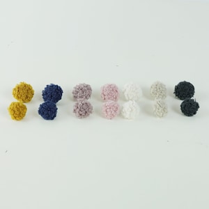 Pode incluir: Um conjunto de 10 pompons fofinhos em várias cores, incluindo amarelo mostarda, azul marinho, rosa pó, rosa claro, branco e cinza. Os pompons estão dispostos em duas filas de cinco.