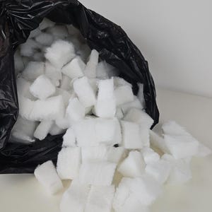 May include: A black plastic bag filled with white, fluffy, square-shaped packing peanuts. The bag is open and the peanuts are spilling out onto a white surface.