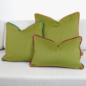May include: Three green velvet throw cushions with different coloured trim. The largest cushion has orange and blue trim, the medium cushion has blue trim, and the smallest cushion has pink and blue trim.