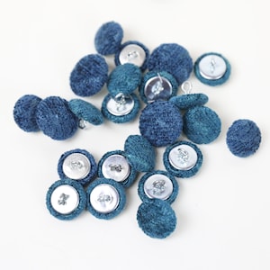 May include: A collection of teal blue fabric covered buttons with silver metal shanks. The buttons are scattered on a white surface.