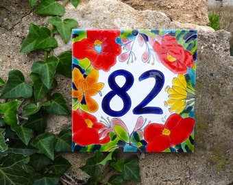 Puede incluir: Un azulejo de cerámica blanco con un diseño floral en tonos rojos, naranjas, amarillos y verdes. El número 82 está impreso en azul en el centro del azulejo.