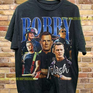 Può includere: T-shirt nera con un collage di immagini e il nome "Bobby" in testo blu stilizzato. La maglietta presenta immagini di uomini in uniforme, con il nome "Nash" in argento. La maglietta è appesa a una gruccia di legno.