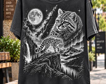 May include: A black short-sleeve t-shirt featuring a detailed white graphic print of a cat, a smaller cat walking on a rocky cliff, a full moon, a starry sky, a waterfall, mountains, and a forest landscape. The shirt has a crew neckline.