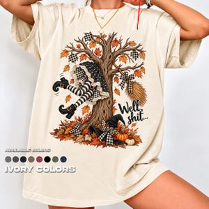 May include: An ivory short-sleeve t-shirt featuring a graphic of a witch upside down in a tree with orange autumn leaves, a broom, a black hat, and pumpkins. The text "Well, shit..." is printed in black script next to the tree trunk.