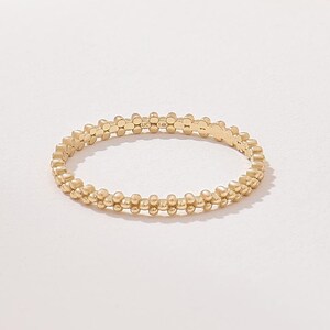 Op de afbeelding: Een dunne gouden ring met een doorlopende band van kleine, uniforme bolvormige kralen gerangschikt in een delicaat, cirkelvormig patroon. Het metaal heeft een gepolijste afwerking die het licht reflecteert op het oppervlak van elke kraal.