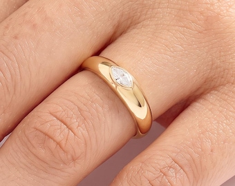 May include: A gold band ring featuring a single marquise-cut clear gemstone set flush into the centre of the polished metal surface. The ring has a smooth, rounded profile and a high-shine finish, showcasing a clean and simple design aesthetic.