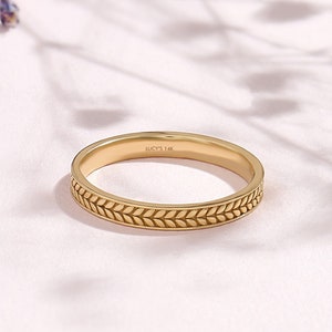 May include: A gold ring with a leaf pattern around the band. The ring is on a pink surface with purple and white flowers in the background. The text "LUCYS 14K" is engraved on the ring.