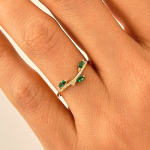 May include: A gold ring featuring a thin, branch-like band adorned with three marquise-cut green stones arranged in a leaf-like pattern. The metal band has a textured finish, and the green stones are set securely along the delicate, curved design.
