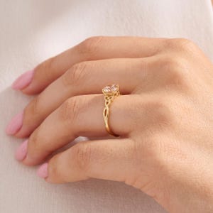 May include: A gold ring featuring a round, clear, faceted gemstone set in a prong setting. The band is crafted with an intricate, openwork knot design that extends from the base of the setting down the sides of the ring, creating a continuous pattern.