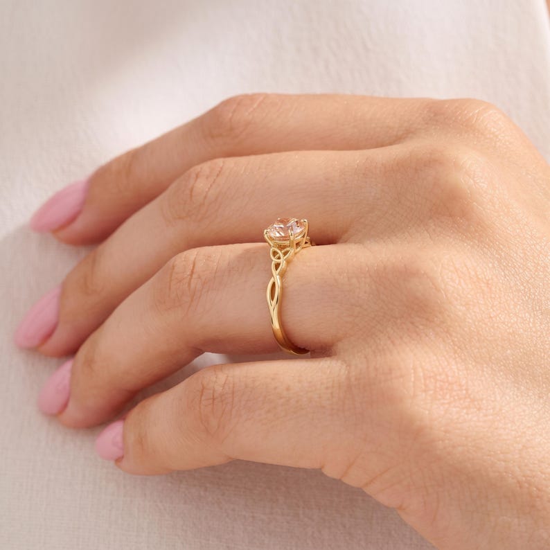 May include: A gold ring featuring a round, clear, faceted gemstone set in a prong setting. The band is crafted with an intricate, openwork knot design that extends from the base of the setting down the sides of the ring, creating a continuous pattern.