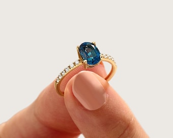 May include: A gold ring featuring a central oval-cut deep blue gemstone held by four prongs. The band is accented with a row of small, clear round-cut stones embedded along each side of the shank, creating a sparkling effect on the yellow gold metal.