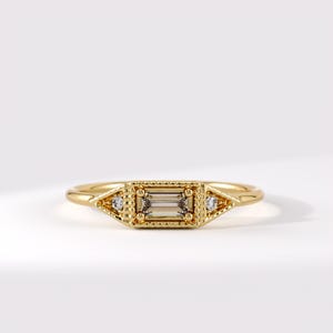 May include: A gold ring featuring a central baguette-cut stone set within a rectangular frame with beaded edges. Two small round stones are positioned on either side of the center setting, integrated into a pointed, textured metal design.