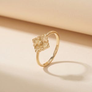 May include: A gold ring with a square floral design. The ring has a slender band and a detailed flower-shaped centrepiece. The ring is made of gold and is set against a neutral background. The ring is a piece of jewellery.