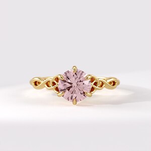 May include: A gold ring featuring a round, pale pink gemstone held by six prongs. The band is crafted with an intricate, interwoven knot design that extends from the central setting, showcasing a polished metallic finish and a symmetrical structure.