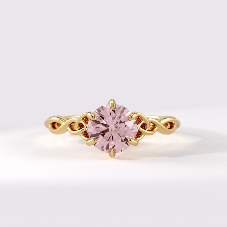 May include: A gold ring featuring a round, pale pink gemstone held by six prongs. The band is crafted with an intricate, interwoven knot design that extends from the central setting, showcasing a polished metallic finish and a symmetrical structure.