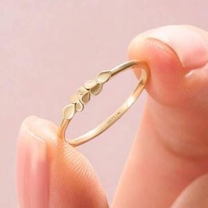 Puede incluir: Un delicado anillo de oro con un diseño floral de pétalos en forma de lágrima. El anillo se muestra sobre un fondo rosa suave. El texto "LUCY'S 14K" está inscrito en la banda del anillo. Una pieza elegante para el uso diario.