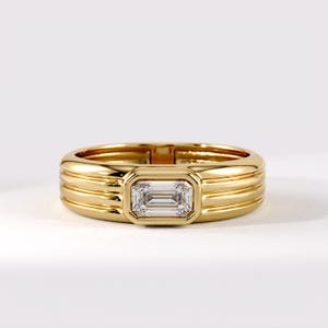 May include: A polished yellow gold ring featuring a horizontal emerald-cut clear gemstone set in a bezel. The band is designed with three distinct horizontal ridges that wrap around the circumference, creating a layered, textured appearance.
