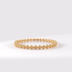 Op de afbeelding: Een goudkleurige ring bestaande uit een doorlopende band van kleine, bolvormige kralen in een herhalend patroon. Het metalen oppervlak heeft een gepolijste, reflecterende afwerking, wat zorgt voor een uniform, cirkelvormig ontwerp rondom de ring.