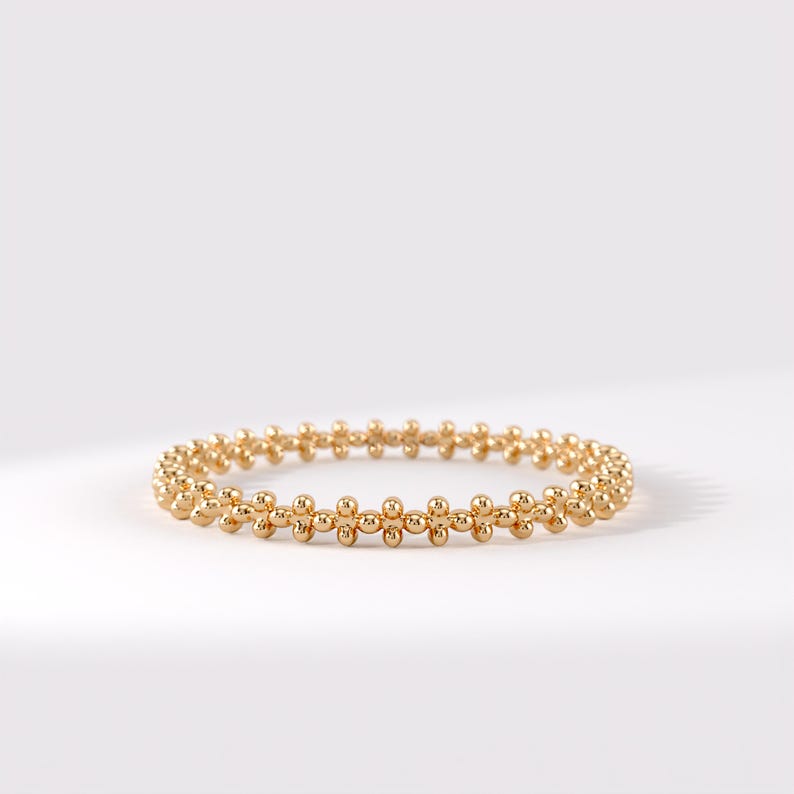 Op de afbeelding: Een goudkleurige ring bestaande uit een doorlopende band van kleine, bolvormige kralen in een herhalend patroon. Het metalen oppervlak heeft een gepolijste, reflecterende afwerking, wat zorgt voor een uniform, cirkelvormig ontwerp rondom de ring.