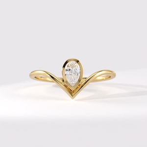 Op de afbeelding: Een gouden V-vormige ring met een enkele peervormig geslepen heldere edelsteen in een gladde zetting. De ringband heeft een gepolijste afwerking en een gebogen, chevron-achtig silhouet dat taps toeloopt naar het midden waar de steen is geplaatst.