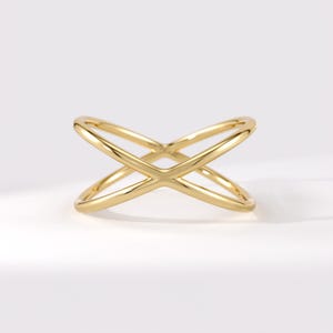 May include: A polished gold ring featuring a smooth, continuous band that crosses over itself in the centre to form an X-shaped design. The metal has a reflective, high-shine finish with rounded edges throughout the entire structure of the ring.