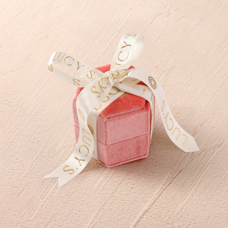 the ring box that products will ben send in is in peachy pink color with creamy colored ribbon tied as a bow.