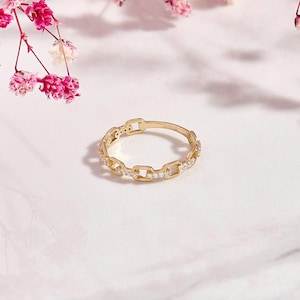 May include: A gold-colored ring with a chain link design. The ring is adorned with small, clear gemstones. The ring is set against a soft, white background with pink flowers in the upper left corner.