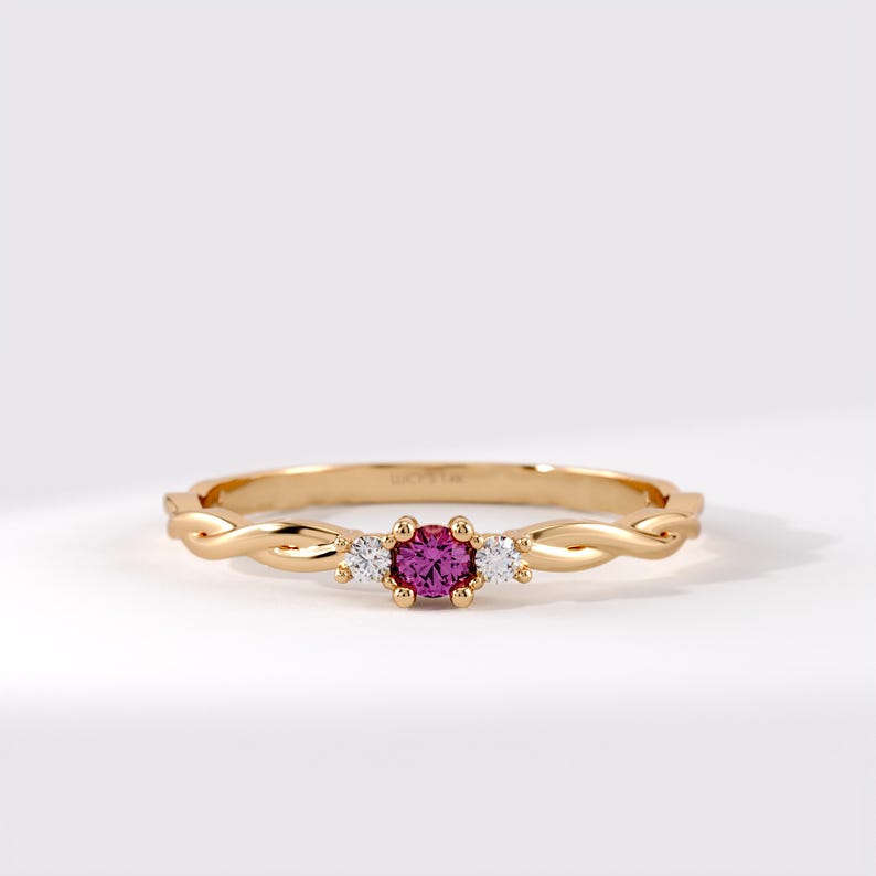 Pode incluir: Um anel de ouro com uma pedra preciosa rosa de corte redondo central ladeada por duas pedras claras menores. A banda tem um design tran&ccedil;ado semelhante a uma corda e est&aacute; gravada com o texto LUCY'S 14K na superf&iacute;cie interna da banda de metal.