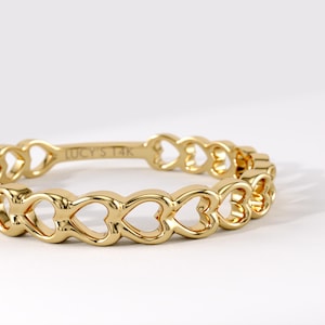May include: A gold ring featuring a continuous band of interconnected heart-shaped cutouts. The interior of the band is clearly engraved with the text LUCY'S 14K, indicating the metal purity. The ring has a polished, reflective finish throughout.
