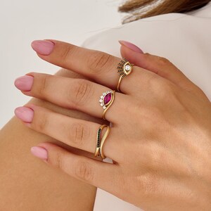 K&ouml;nnte beinhalten: Drei goldene Ringe mit schwarzen und wei&szlig;en Steinen. Der obere Ring hat einen wei&szlig;en Stein mit schwarzen Steinen, die eine Augenform bilden. Der mittlere Ring hat einen roten Stein mit wei&szlig;en Steinen, die eine Kronenform bilden. Der untere Ring hat ein gebogenes Band mit schwarzen Steinen.