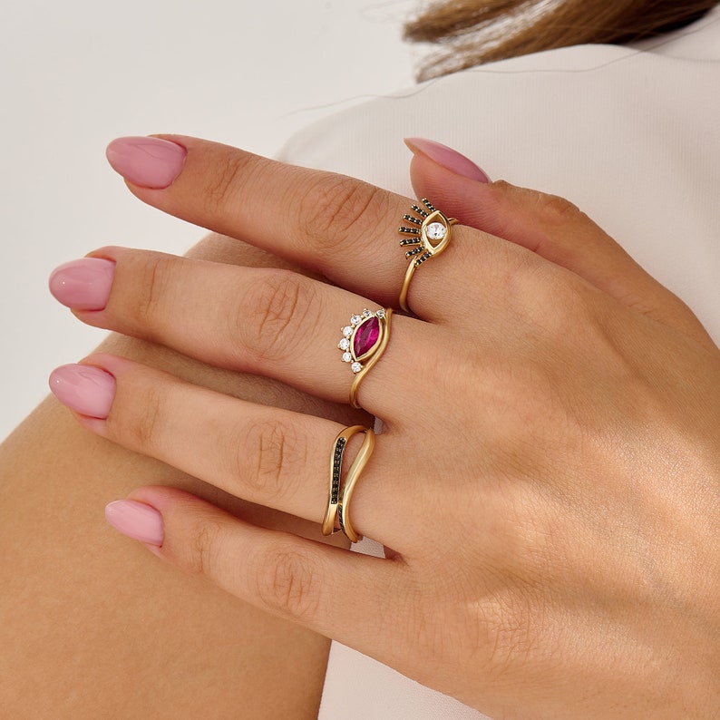 K&ouml;nnte beinhalten: Drei goldene Ringe mit schwarzen und wei&szlig;en Steinen. Der obere Ring hat einen wei&szlig;en Stein mit schwarzen Steinen, die eine Augenform bilden. Der mittlere Ring hat einen roten Stein mit wei&szlig;en Steinen, die eine Kronenform bilden. Der untere Ring hat ein gebogenes Band mit schwarzen Steinen.