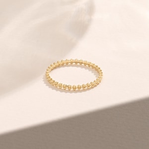 以下が含まれることがあります： テクスチャ加工されたビーズデザインのゴールドリング。リングは円形で、バンド全体に小さく均等に配置されたビーズが特徴です。リングは、柔らかいオフホワイトの背景に配置され、微妙な影があります。
