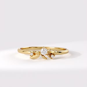 May include: A gold ring featuring a central round clear gemstone flanked by two smaller round clear gemstones, all set within a delicate metal leaf motif. The band is thin and polished, showcasing a smooth, continuous circular design.